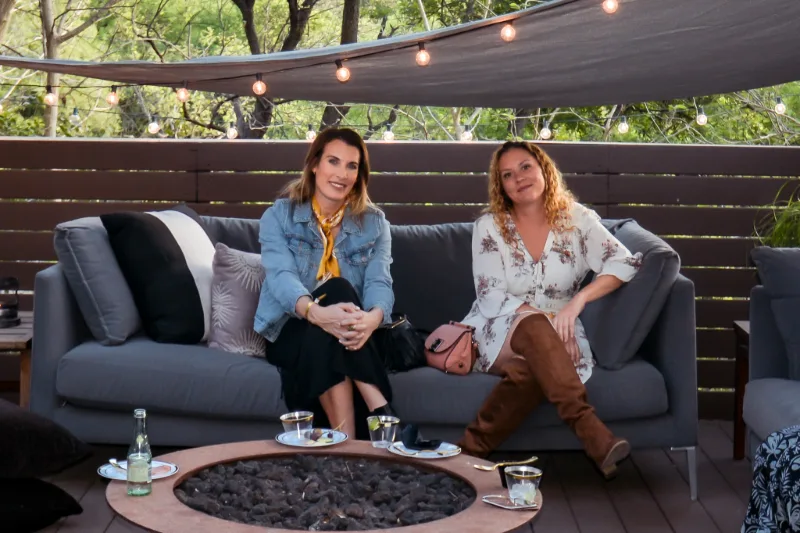 Two people sitting on an outdoor sofa beside a fire pit under string lights.