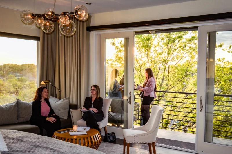 Two people talking on a sofa and an armchair indoors while others stand on the balcony outside.