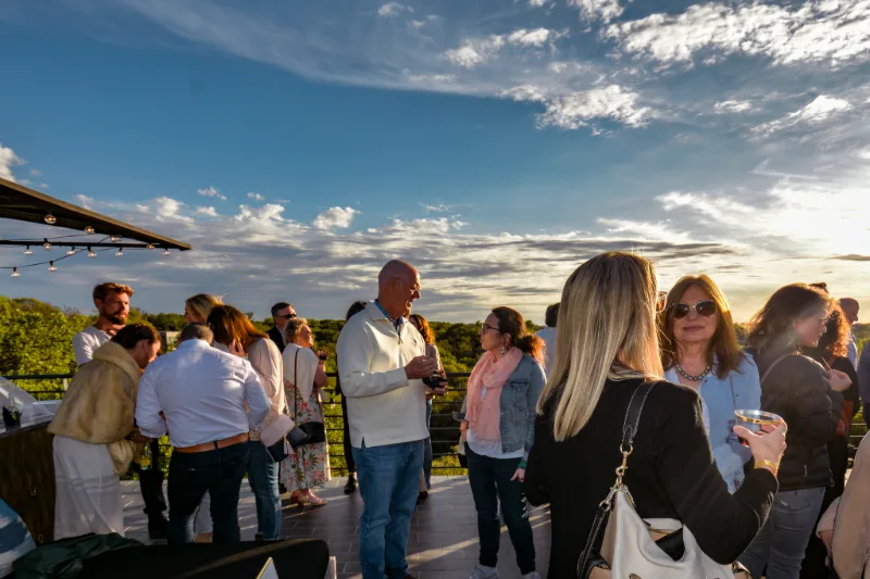 People mingling on a rooftop terrace under a blue sky, with string lights and treetops beyond.