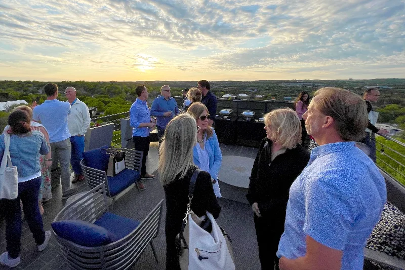 People talking in small groups on a rooftop terrace as the sun sets over the trees.